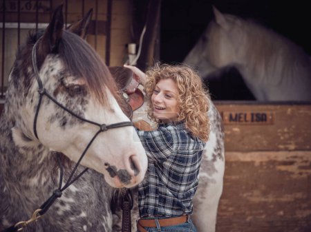 Foto de Jinete femenino positivo en camisa a cuadros de pie cerca de lindo caballo con brida en establo con puesto de madera en el área rural - Imagen libre de derechos