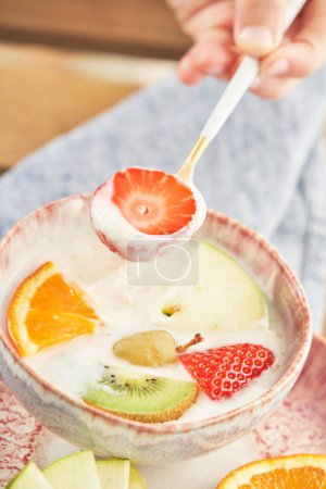 Téléchargez les photos : Main de la culture personne méconnaissable tenant cuillère avec des tranches de fraise sur le bol avec du yaourt savoureux et des fruits pendant le petit déjeuner dans la cuisine - en image libre de droit