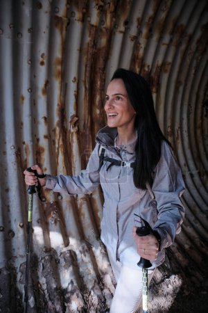 Photo for Cheerful Hispanic female with black hair and trekking poles standing in old neglected tunnel with shabby rusted walls during trip - Royalty Free Image