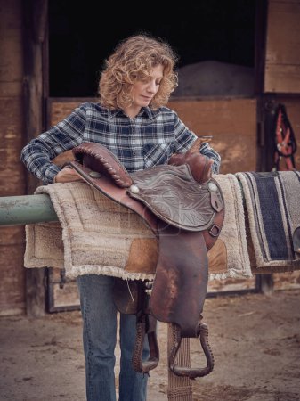 Foto de Agricultora en camisa a cuadros que pone silla de montar occidental en barandilla de metal con almohadillas cerca de establo de mala calidad en el área rural en el día de verano - Imagen libre de derechos