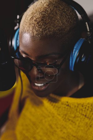 Photo for From above of positive young African American female singer with short hair wearing eyeglasses and headphones while recording song via microphone in studio - Royalty Free Image