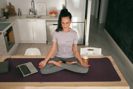 Photo for Full body of peaceful Hispanic female with closed eyes sitting in Lotus pose with beer on table with netbook during yoga lesson in kitchen - Royalty Free Image