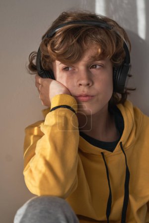 Foto de Niño reflexivo con auriculares y suéter amarillo escuchando canciones mientras se relaja solo en casa y mirando hacia otro lado - Imagen libre de derechos