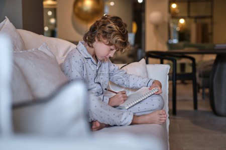 Photo for Side view of focused preteen child with barefoot crossed legs sitting on sofa and drawing making pencil sketch in living room - Royalty Free Image