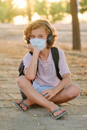 Photo for Full body of child in protective mask sitting on pathway in park and leaning on hand while listening to music in headphones during COVID epidemic - Royalty Free Image