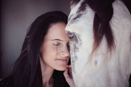 Foto de Amable propietaria femenina con ojos cerrados acariciando bozal de caballo con mancha blanca de pie en establo en zona rural - Imagen libre de derechos