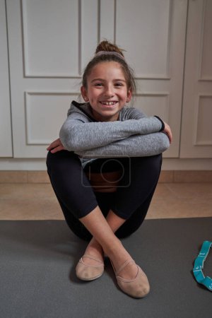 Photo for Full body portrait of positive preteen gymnast in sportswear smiling and sitting on exercise mat at home during gymnastics training - Royalty Free Image