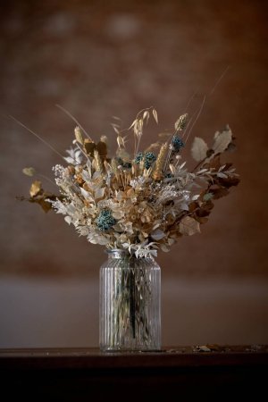 Foto de Ramo de flores secas con ramitas colocadas en jarrón de vidrio en la mesa contra el fondo borroso en la habitación de estilo antiguo - Imagen libre de derechos