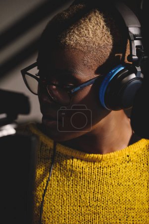 Foto de Retrato de cantante femenina afroamericana seria en gafas y auriculares grabando canción cerca del micrófono en el estudio mientras mira hacia otro lado - Imagen libre de derechos
