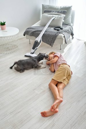 Photo for High angle of cute Schnauzer dog licking face of exhausted boy resting on floor near vacuum cleaner after doing household chores - Royalty Free Image