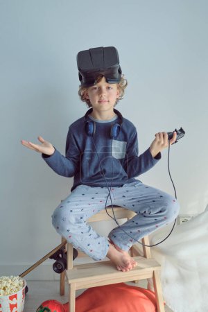 Foto de Cuerpo completo de lindo niño preadolescente que usa ropa de dormir con auriculares de realidad virtual en la cabeza y auriculares en el cuello con joystick sentado en la escalera de taburete - Imagen libre de derechos
