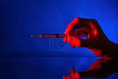Photo for Crop person holding medical syringe with needle filled with COVID 19 vaccine on reflective surface in dark room - Royalty Free Image