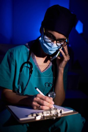 Foto de Médico femenino concentrado en uniforme y máscara sentado en escaleras con portapapeles y tomando notas en la historia clínica mientras habla en smartphone en pasillo con luz de neón - Imagen libre de derechos