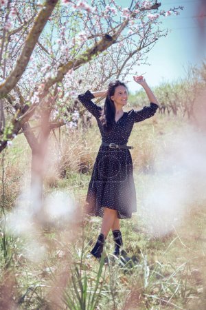 Photo for Full body of positive Hispanic woman looking up with raised arms while standing near blooming almond trees on summer day - Royalty Free Image