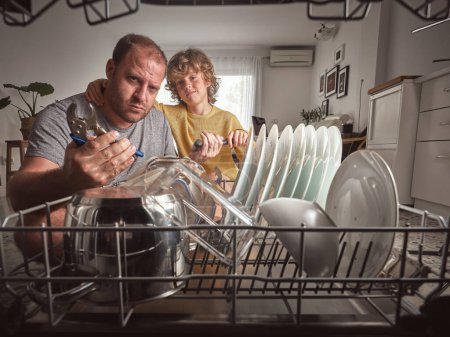Téléchargez les photos : Père confiant avec pince de verrouillage spéciale et fils avec marteau regardant la caméra près du lave-vaisselle moderne cassé dans la cuisine légère - en image libre de droit