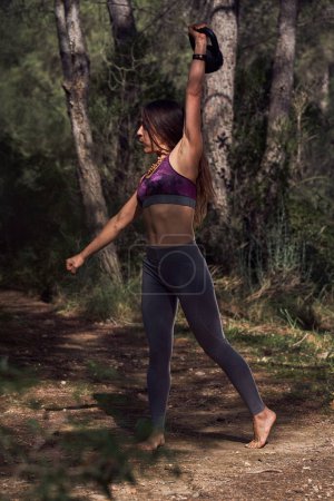 Téléchargez les photos : Vue latérale complète du corps d'une femme forte en tenue de sport soulevant une lourde kettlebell dans un bras levé pendant l'entraînement dans les bois - en image libre de droit