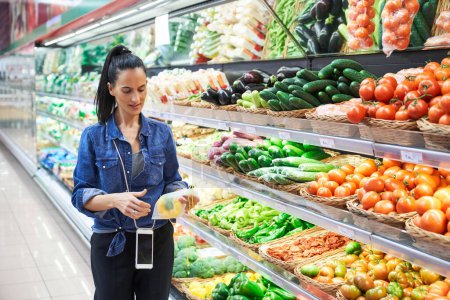 Foto de Señora étnica de pie cerca de los estantes con una variedad de verduras en la tienda de comestibles y la compra de pimienta fresca en bolsa de plástico - Imagen libre de derechos