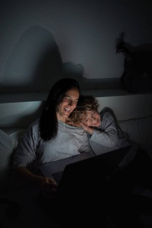 Téléchargez les photos : Mère ravie avec enfant en tenue décontractée riant et surfant netbook tout en faisant des devoirs ensemble sur le lit dans la chambre sombre - en image libre de droit