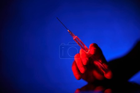 Foto de Primer plano de la persona irreconocible que sostiene la jeringa médica con aguja llena de vacuna contra el coronavirus en habitación oscura sobre fondo azul - Imagen libre de derechos