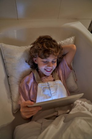 Téléchargez les photos : De dessus de sourire enfant avec les cheveux bouclés reposant dans la baignoire avec oreiller et couverture et regarder des vidéos sur tablette dans la salle de bain à la maison - en image libre de droit