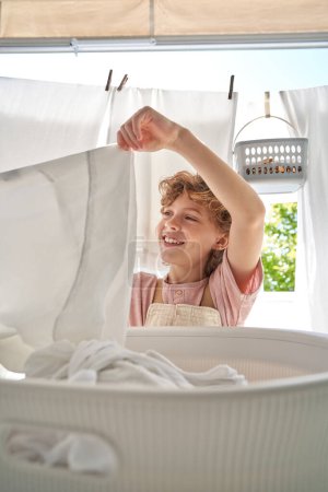 Photo for Smiling child standing under clothesline and taking white sheets from laundry basin while doing household routine on balcony at home - Royalty Free Image