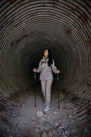 Photo for Full body of Hispanic female with trekking poles looking up while observing shabby rough wall in long old tunnel during trip - Royalty Free Image