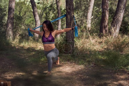 Téléchargez les photos : Corps entier de femme hispanique pieds nus concentrée faisant de l'exercice de flexion tout en tirant une corde de résistance pour maintenir l'équilibre et renforcer les muscles pendant l'entraînement de fitness en forêt - en image libre de droit