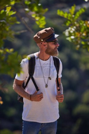 Téléchargez les photos : Touriste masculin barbu dans des lunettes de soleil et casquette avec sac à dos debout près des arbres verts contre la pente de la montagne et regardant loin - en image libre de droit