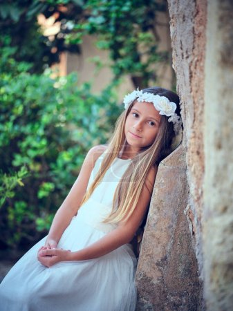 Téléchargez les photos : Femme calme en robe blanche et couronne florale regardant la caméra tout en s'appuyant sur le mur dans un parc verdoyant - en image libre de droit