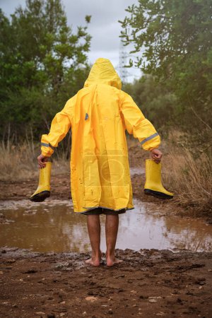 Téléchargez les photos : Vue arrière d'un enfant méconnaissable en imperméable jaune debout sur un sol salissant près de la flaque d'eau tout en explorant les bois le jour de pluie - en image libre de droit