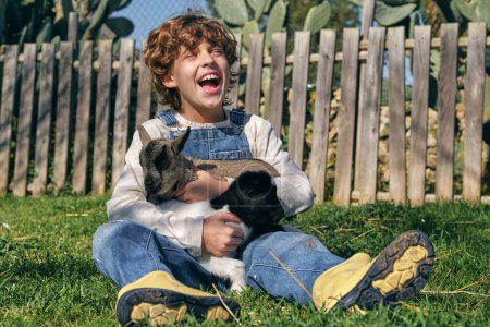Photo for Full body of cheerful kid with blond curly hair in denim overall sitting on grassy meadow and laughing while stroking cute fluffy goatlings in countryside - Royalty Free Image