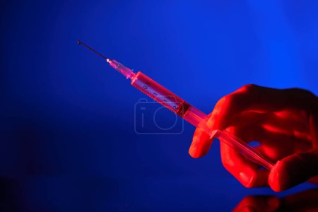 Photo for Crop anonymous person holding medical syringe with needle filled with COVID19 vaccine in neon lighting in dark room - Royalty Free Image