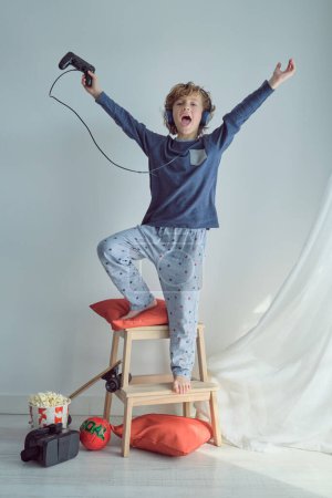 Photo for Full body of cheerful barefoot boy in pajama and headphones raising arms and screaming happily while playing video game with game pad and celebrating victory against white wall - Royalty Free Image