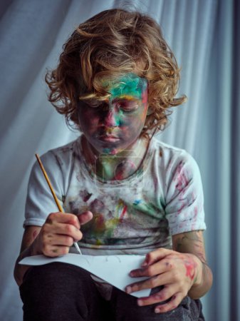 Foto de Niño creativo concentrado cubierto de pintura con camiseta manchada y pantalones que sostienen el dibujo del pincel sobre papel colocado sobre las rodillas - Imagen libre de derechos