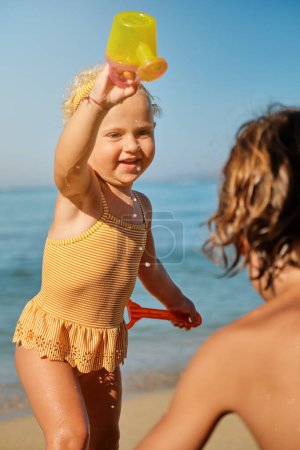 Téléchargez les photos : Mignon enfant en maillot de bain jaune s'amuser avec arrosoir et regarder garçon près de la mer sur la plage de sable - en image libre de droit