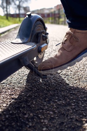 Foto de Crop hombre anónimo en calzado de cuero perforado estacionamiento scooter eléctrico en camino de asfalto en la calle durante el día - Imagen libre de derechos