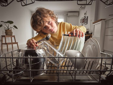 Photo for Focused boy with special instruments in hands checking broken dishwasher with dishware on racks while standing in light kitchen at home - Royalty Free Image
