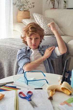 Photo for Displeased preteen boy showing muscles and frowning while sitting at table with medical mask and stationery and doing homework in living room at home during COVID 19 epidemic - Royalty Free Image