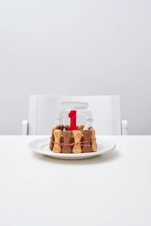Foto de Apetitivo pastel casero decorado con golosinas de hueso y vela roja en plato blanco colocado cerca de la silla vacía en la mesa gris - Imagen libre de derechos