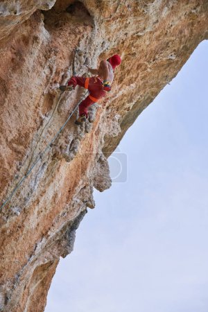 Photo for From below back view of unrecognizable alpinist in red clothes climbing on edge of cliff with rope and carbines - Royalty Free Image