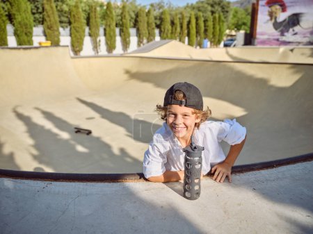 Photo for Cheerful kid with bottle of water looking at camera and leaning on ramp in skate park on sunny day - Royalty Free Image