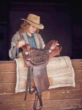 Foto de Mujer rubia rizada enfocada en la ropa de vaquera mirando hacia abajo mientras pone la silla de montar en la alfombra colocada en el establo de madera - Imagen libre de derechos