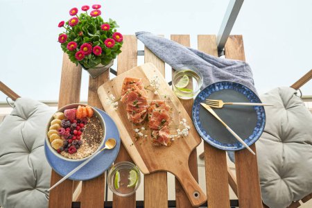 Photo for Top view of yummy toasts with prosciutto served on cutting board near bowl of healthy oatmeal with assorted berries and lime water glasses placed on table near houseplant in restaurant - Royalty Free Image