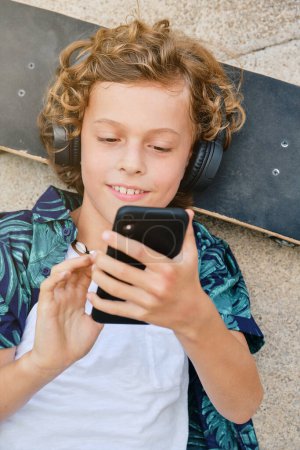 Téléchargez les photos : Vue du dessus du garçon souriant écoutant de la musique dans les écouteurs et surfant sur les médias sociaux sur le téléphone mobile tout en étant allongé sur le skateboard sur la rue de la ville - en image libre de droit