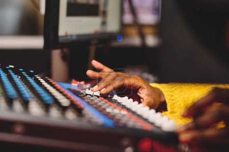 Téléchargez les photos : Femme afro-américaine de culture méconnaissable en pull jaune à l'aide d'un panneau de mélange avec boutons de commande tout en travaillant dans un studio de radiodiffusion - en image libre de droit