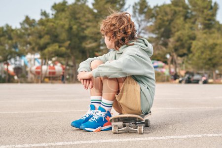 Photo for Side view of anonymous teen boy in cool outfit sitting on skateboard and embracing knees in street - Royalty Free Image