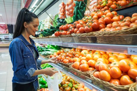 Photo for Side view of content female customer with nylon bag choosing fresh tomatoes from stall during grocery shopping in light supermarket - Royalty Free Image