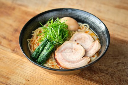 High angle of ceramic bowl of ramen soup with pork meat herbs vegetables and boiled egg on wooden table against blurred background