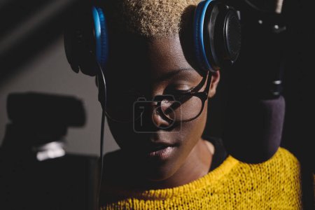 Crop portrait of concentrated young ethnic female radio host in glasses and headphones raised on forehead near microphone in dark studio looking down
