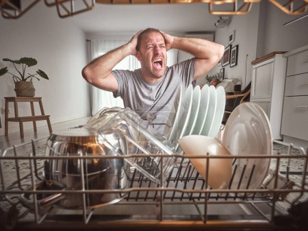 Foto de Hombre molesto gritando con las manos en la cabeza cerca de lavavajillas abierto moderno con varios utensilios de cocina durante la rutina del hogar en la cocina ligera - Imagen libre de derechos